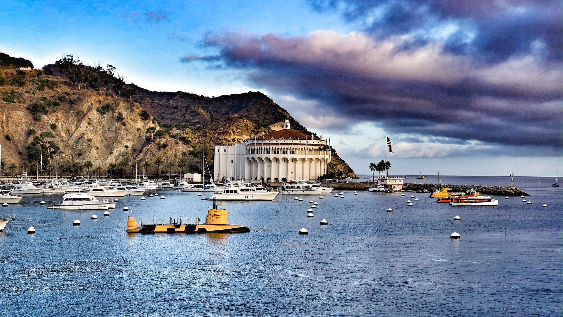 A far view of a circular building, mountains, the ocean with boats, and clouds